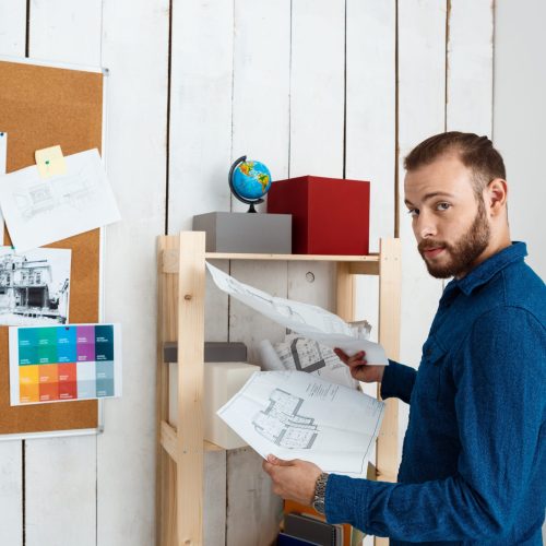 Young successful architector smiling, holding drawings, standing in office.